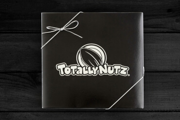 black square box with silver totally nutz logo embossed on top and closed with a silver ribbon, box has gingham print bottom and the photo is taken on a white wood background, all occasion gift box 
